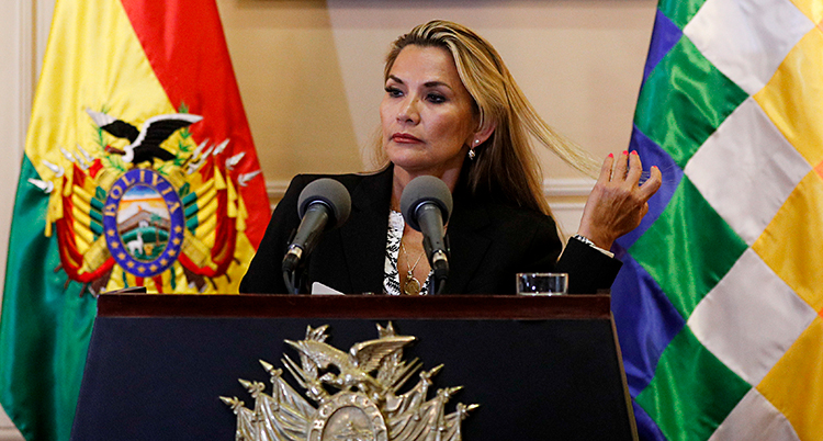Jeanine Áñez står i en talarstol och pratar. Hon har långt blont hår och en svart kavaj. I bakgrunden syns flaggor, bland annat den bolivianska flaggan.