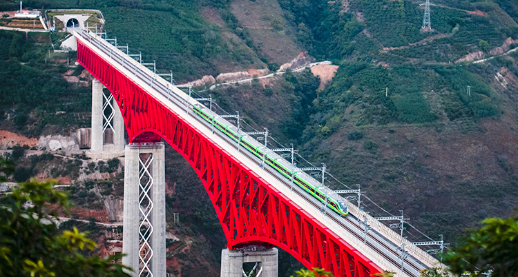 En bro i rött över en stor dal och en flod, fotad från luften. Ett tåg kör över bron.