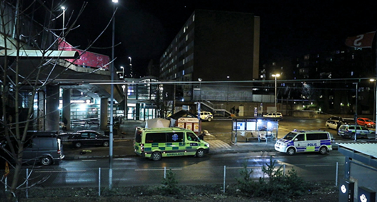 Det är mörkt ute. En polisbil och en ambulans står vid ett torg.