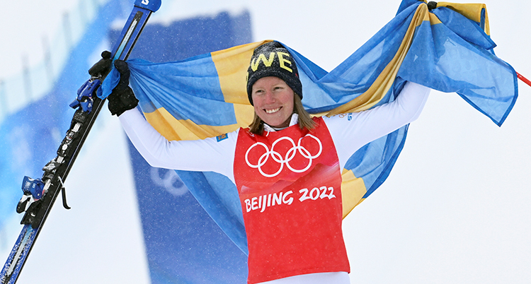 Hon sträcker upp armarna i luften och ler. Hon håller en svensk flagga och sina skidor.