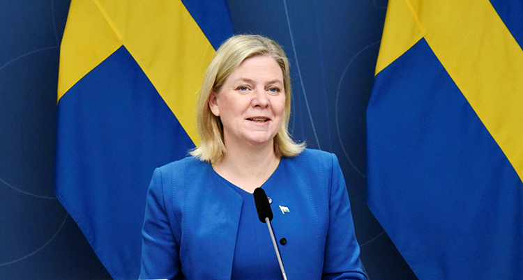 Hon har blå kavaj och ljust hår. Hon står framför svenska flaggor och pratar i en mikrofon.
