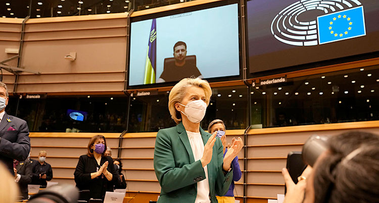 Hon står upp och applåderar. Hon har munskydd. Hon står i en stor sal i EUs byggnad. På en stor skärm bakom henne syns Ukrainas president.
