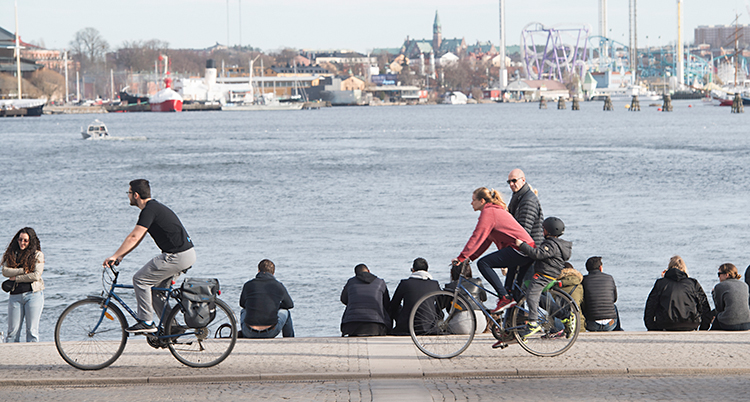 En bild från Nybrokajen i Stockholm. Människor cyklar, promenerar, sitter och umgås, på trapporna vid vattnet.