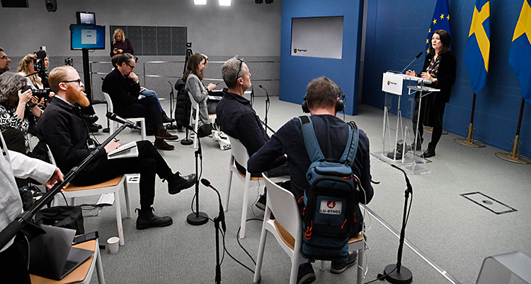 Hon står i ett rum och pratar i en mikrofon framför journalister som sitter på stolar.