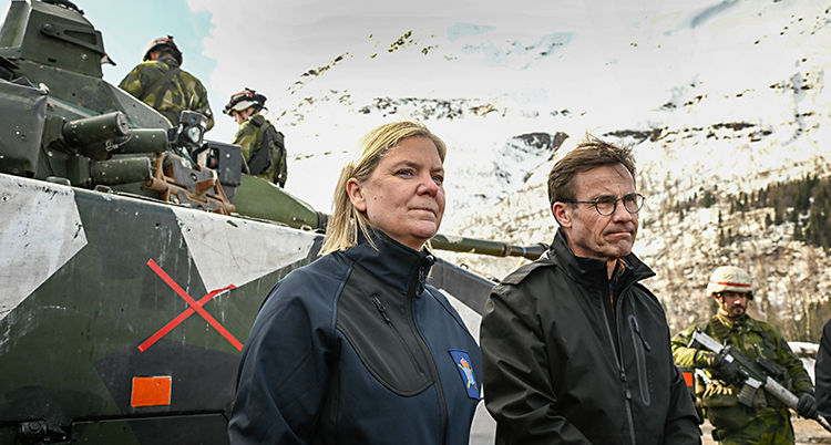 Magdalena och Ulf är utomhus. De står framför en stridsvagn. Runt dem står militärer.