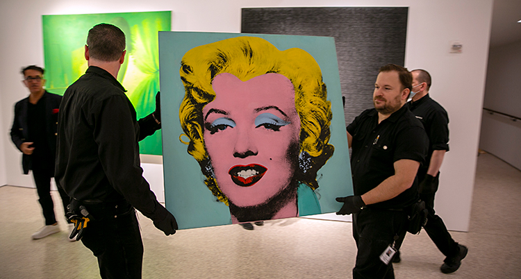 Två män bär på en tavla som visar Marilyn Monroe i färgerna blått, gult och rosa.