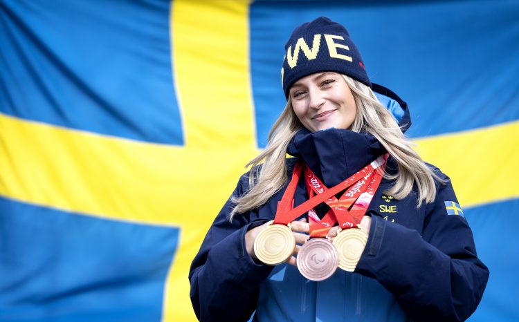 Tjej med mössa framför svenska flaggan med flera olympiska medaljer runt halsen.
