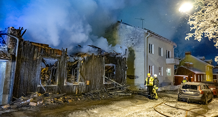 Ett lågt gammalt hus av trä är helt förstört. Det ryker från taket. Intill syns ett vitt, högre hus.