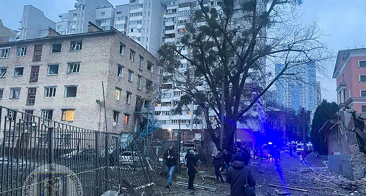 En stad med stora trasiga hus. Ett träd syns. Människor står på gatan och tittar på det som är förstört.