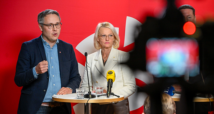 De står bredvid varandra vid ett bord. Det är mikrofoner på bordet. I förgrunden syns en kamera.