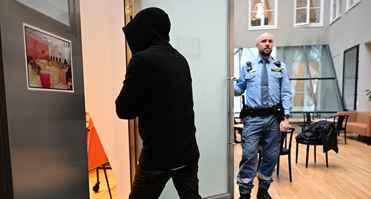 En person i mörk jacka med luva går in genom en dörr. En vakt står vid dörren.