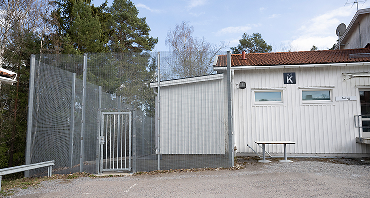 Ett vitt hus. Bredvid huset finns ett högt stängsel med en dörr av metall.