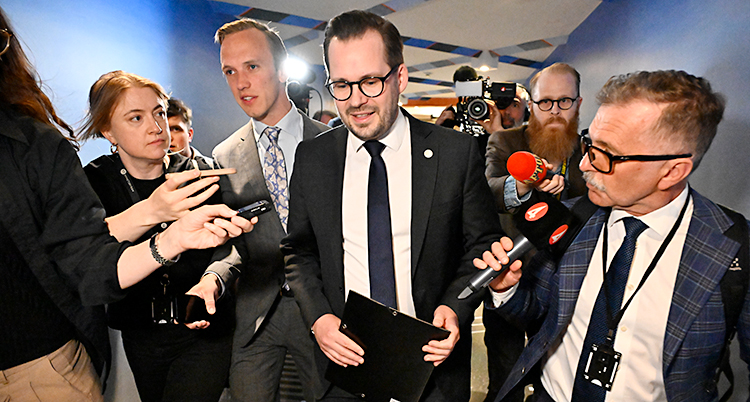 En man kostym går genom en korridor. Journalister med mikrofoner går bredvid honom.
