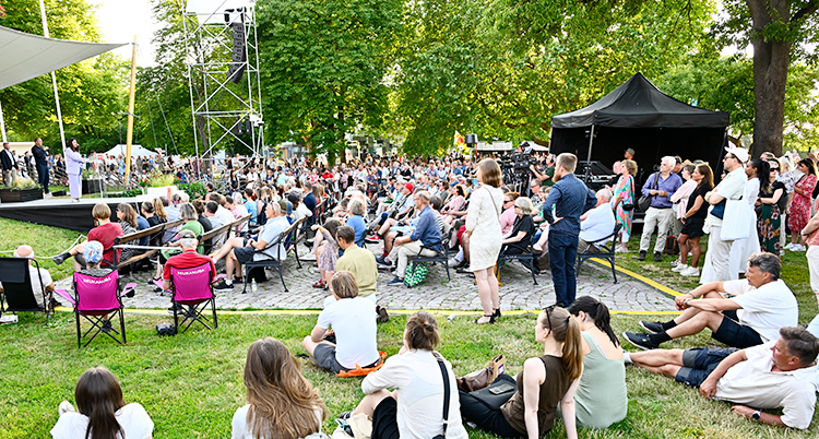 Folk är utomhus i en park och lyssnar på en person som står och talar på en scen.