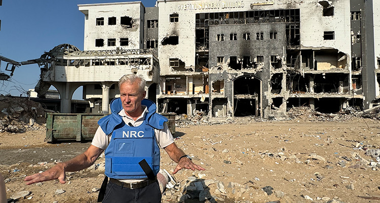 Han står i blå skyddsväst framför ett bombat hus.