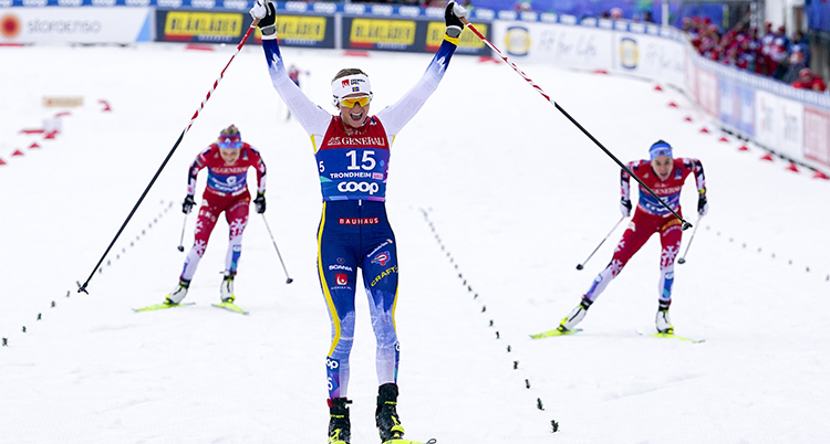Svensk skidåkare åker över mållinjen och sträcker armarna i luften. I bakgrunden syns två norska skidåkare.