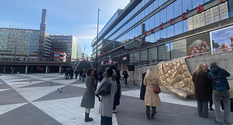 Konstverket finns på en mur. Torget syns bakom. Människor står på torget.